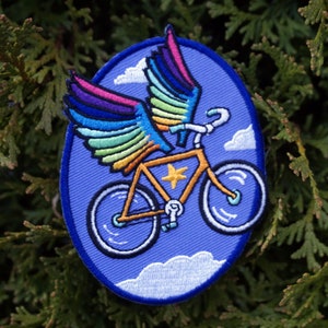 May include: A blue embroidered patch featuring a yellow bicycle with rainbow wings flying through the air. The bicycle has a star on the frame and is surrounded by white clouds.