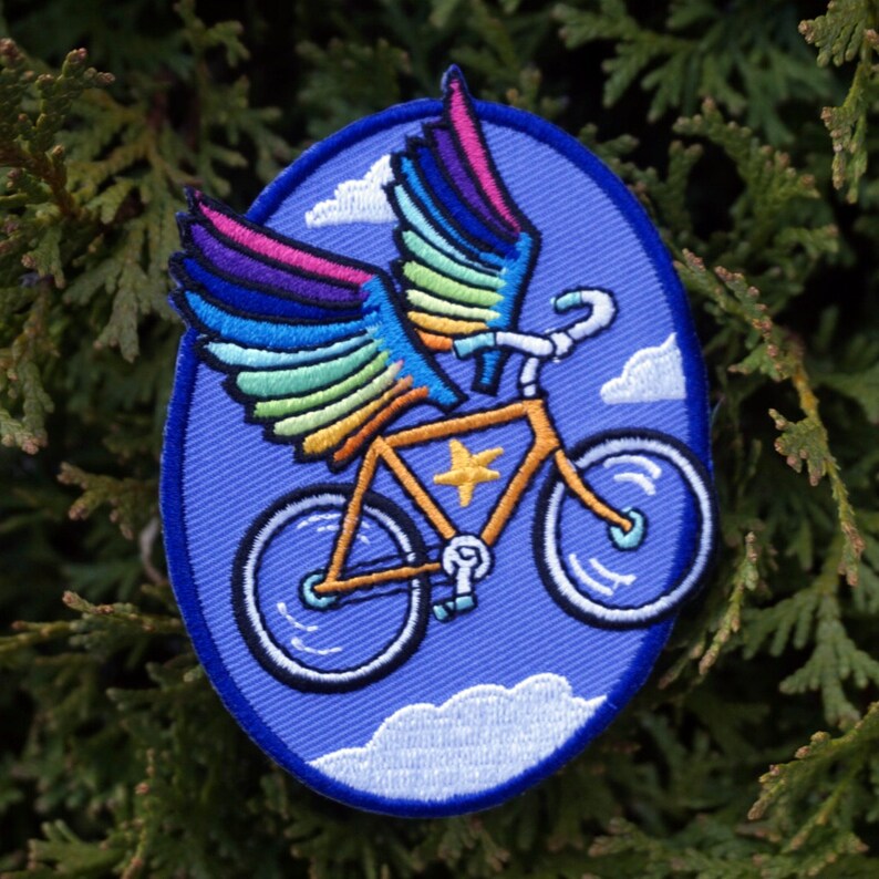 May include: A blue embroidered patch featuring a yellow bicycle with rainbow wings flying through the air. The bicycle has a star on the frame and is surrounded by white clouds.