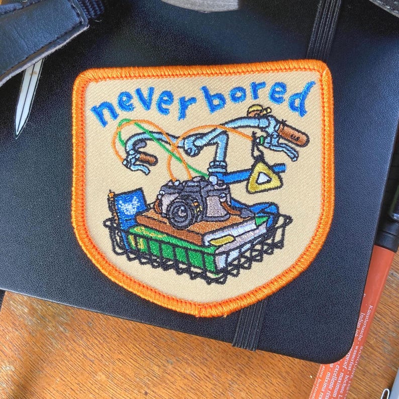 May include: Embroidered patch with the words "never bored" in blue script. The patch features a bicycle handlebar, camera, books, and a triangle. The patch has a tan background with an orange border.