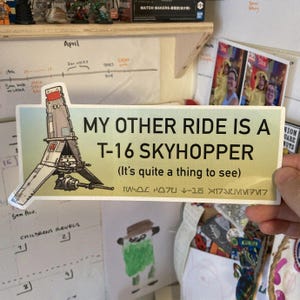 Peut inclure: Un autocollant jaune et blanc avec une illustration de dessin animé d'un vaisseau spatial avec le texte "My other ride is a T-16 Skyhopper (It's quite a thing to see)"