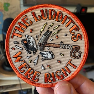 May include: Circular embroidered patch with the text "THE LUDDITES WERE RIGHT" in orange thread. The patch depicts a hammer smashing machinery, with lightning bolts and debris. It has an orange border and a beige background.