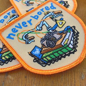 May include: Embroidered patches with the words "never bored" in blue script. The patches feature a colourful illustration of a bicycle with orange, green, and blue accents. The patches have an orange border and a beige background.