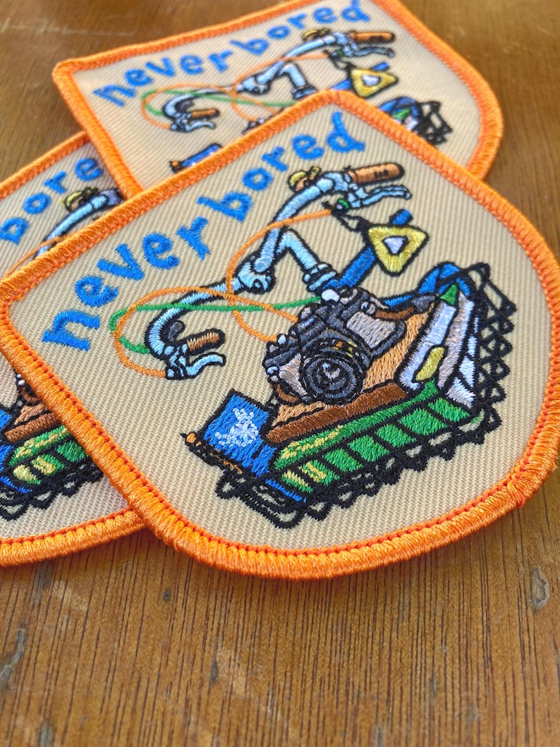 May include: Embroidered patches with the words "never bored" in blue script. The patches feature a colourful illustration of a bicycle with orange, green, and blue accents. The patches have an orange border and a beige background.