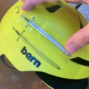 May include: Bright yellow bicycle helmet with two sword decals and the word "bern" in navy blue. The helmet has a black visor and a black strap. The helmet is being held by a person.