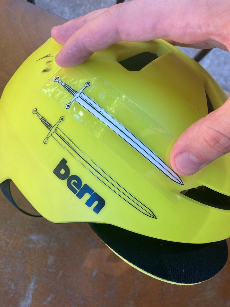 May include: Bright yellow bicycle helmet with two sword decals and the word "bern" in navy blue. The helmet has a black visor and a black strap. The helmet is being held by a person.