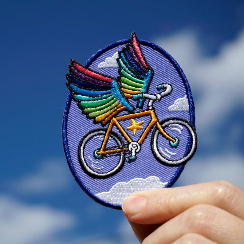 May include: A purple and blue embroidered patch of a bicycle with rainbow wings flying in a blue sky with white clouds.