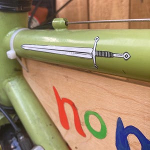 May include: A light green bicycle frame with a white sword decal. The wooden panel attached to the frame has the word "hon" in red, green, and blue. The bike has a silver bell and black tires.