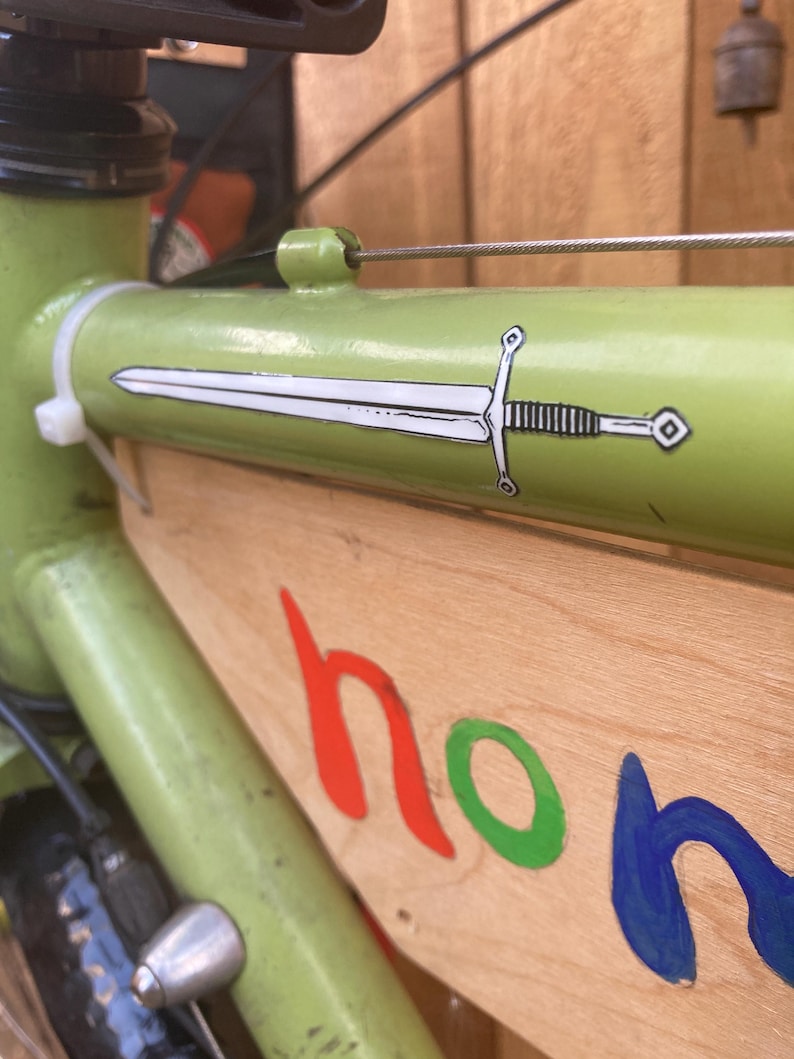 May include: A light green bicycle frame with a white sword decal. The wooden panel attached to the frame has the word "hon" in red, green, and blue. The bike has a silver bell and black tires.