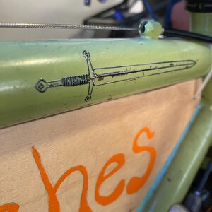 May include: Close-up of a green bicycle frame with a sword graphic. The sword is black and white, and the frame has a distressed look. The wooden panel below has orange lettering.