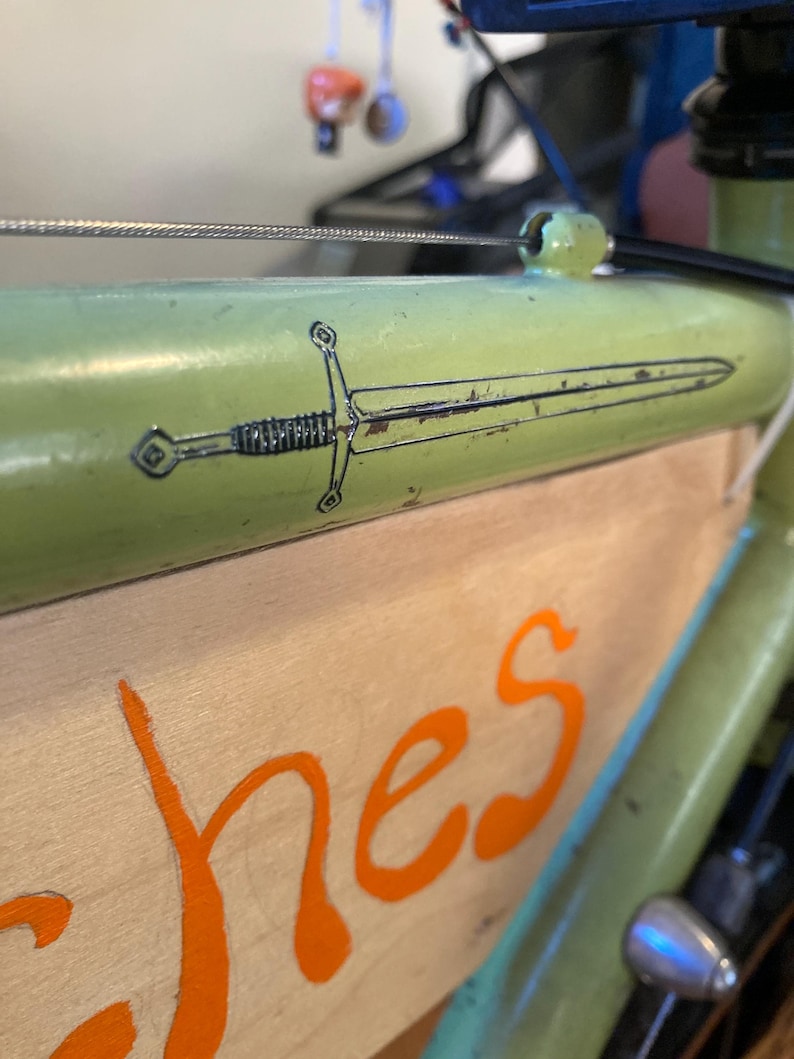 May include: Close-up of a green bicycle frame with a sword graphic. The sword is black and white, and the frame has a distressed look. The wooden panel below has orange lettering.