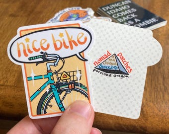 May include: A sticker with a cartoon illustration of a bicycle with the text "nice bike" in a speech bubble. The sticker also includes a logo with the text "Nomad and Stitched Since 2015 Patches Portland Oregon".