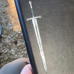 May include: A silver sword sticker with a detailed hilt and blade is affixed to a black, textured surface. The sticker has a metallic sheen, reflecting light. The sword design is a classic, medieval style.