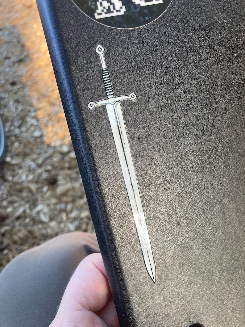 May include: A silver sword sticker with a detailed hilt and blade is affixed to a black, textured surface. The sticker has a metallic sheen, reflecting light. The sword design is a classic, medieval style.