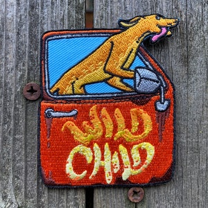 May include: Embroidered patch featuring a cartoon dog in a car, with the text "WILD CHILD" in yellow and orange. The patch has a blue background and an orange car door. The dog has a pink tongue.