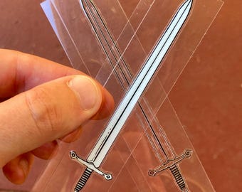 May include: Two transparent stickers featuring a black and white sword design. The swords are crossed, with detailed hilts and blades. The stickers are held against a neutral background.