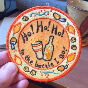 May include: Embroidered patch with a circular design. The patch features the text "Ho! Ho! Ho!" above an illustration of a glass and bottle. The phrase "to the bottle I go" is written below. The patch has an orange and yellow color scheme.
