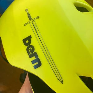May include: Bright yellow helmet with the word "bern" in black and a black sword graphic. The helmet has several ventilation holes and a black strap. The helmet is designed for sports.