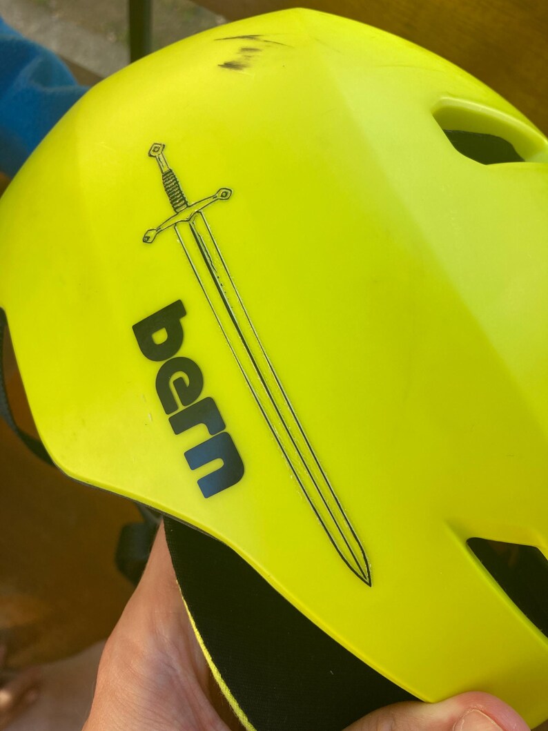 May include: Bright yellow helmet with the word "bern" in black and a black sword graphic. The helmet has several ventilation holes and a black strap. The helmet is designed for sports.