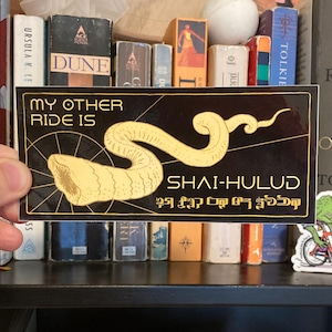 May include: Black sticker with gold text and a stylized sandworm illustration. The text reads "MY OTHER RIDE IS SHAI-HULUD" with additional text below. The sticker is rectangular and has a black background.