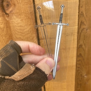 May include: Two transparent stickers featuring a sword design. One sticker has a black outline, while the other has a white blade. The swords have detailed hilts and crossguards, perfect for fantasy enthusiasts.