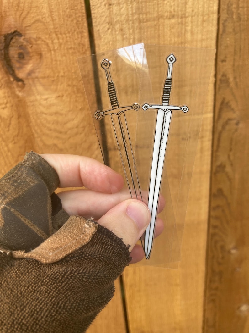 May include: Two transparent stickers featuring a sword design. One sticker has a black outline, while the other has a white blade. The swords have detailed hilts and crossguards, perfect for fantasy enthusiasts.