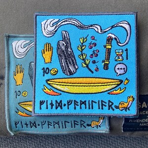 Puede incluir: Dos parches bordados. El parche más grande es cuadrado con un fondo azul claro y presenta varios símbolos en amarillo, negro y blanco. El parche más pequeño tiene un diseño similar con un fondo verde azulado. Ambos parches tienen texto en la parte inferior.