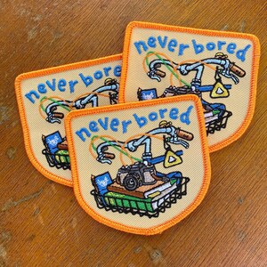 May include: Three embroidered patches with a bicycle design. Each patch features a bicycle with the words "never bored" in blue. The bicycle has a camera, books, and a basket. The patches have a yellow background with an orange border.