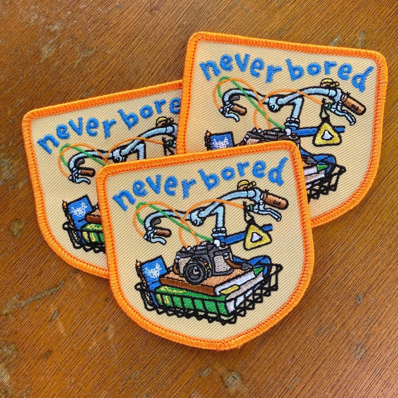 May include: Three embroidered patches with a bicycle design. Each patch features a bicycle with the words "never bored" in blue. The bicycle has a camera, books, and a basket. The patches have a yellow background with an orange border.