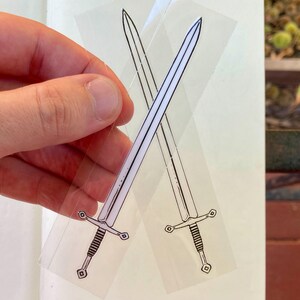 May include: Two transparent stickers featuring crossed swords. Each sword is depicted in black and white, with a detailed hilt and blade. The stickers are held against a white background, with a hand visible.