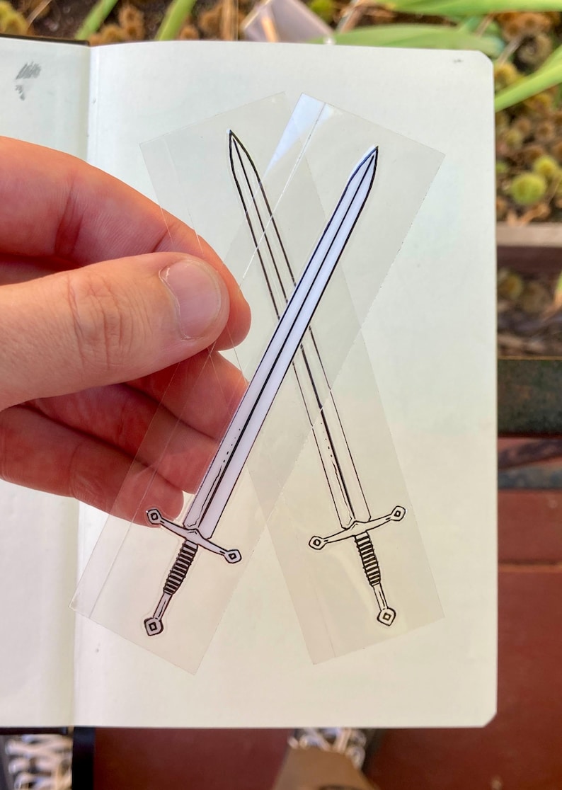 May include: Two transparent stickers featuring crossed swords. Each sword is depicted in black and white, with a detailed hilt and blade. The stickers are held against a white background, with a hand visible.
