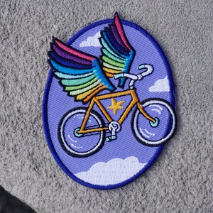 May include: An embroidered patch featuring a bicycle with rainbow wings flying through clouds. The bicycle is yellow with a white star on the frame. The patch is on a purple background.