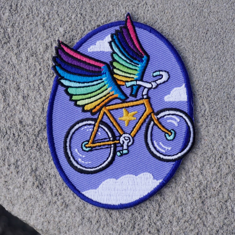 May include: An embroidered patch featuring a bicycle with rainbow wings flying through clouds. The bicycle is yellow with a white star on the frame. The patch is on a purple background.