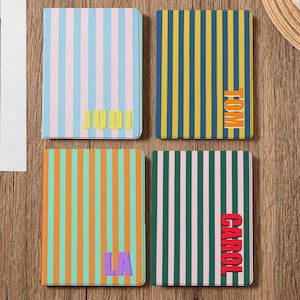 May include: Four notebooks with vertical striped patterns in various colors: blue, pink, yellow, green, and orange. Each notebook has a different name printed on it: JUDI, TOM, LA, and CAROL. A cup of coffee and a straw mat are in the background.