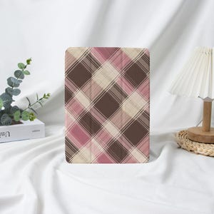 May include: A tablet case featuring a plaid design in brown, pink, and beige. The rectangular case has a folding cover. The pattern consists of intersecting lines, forming a grid. The case is displayed on a white surface, with a lamp and greenery in the background.