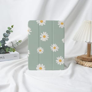 May include: A pale green tablet case adorned with a daisy pattern. The case has a folding cover and features white daisies with yellow centres. The case is designed to protect a tablet.