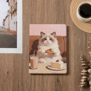 May include: A tablet case featuring a fluffy white cat with blue eyes, holding a slice of pizza. A glass of pink drink and a slice of cake are on the table. The background is a soft pink.