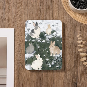 以下が含まれることがあります： 雪原にいるウサギのイラストが描かれた長方形の装飾品。ウサギはグレー、ブラウン、白の色合いで描かれ、緑の葉と白い雪を背景にしています。全体的な美しさは柔らかく、魅力的です。