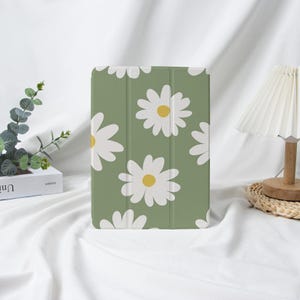 Può includere: Una custodia verde per tablet con un motivo ripetuto di margherite bianche con centri gialli. La custodia è in piedi su una superficie bianca. Il design è semplice e floreale.