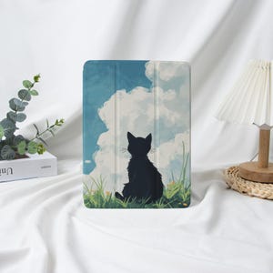 以下が含まれることがあります： 青い空と白い雲を背景に、草むらに座る黒猫のシルエットが描かれたiPadケース。ケースは折りたたみ式のカバーと滑らかな仕上がりで、保護とスタイルを提供します。