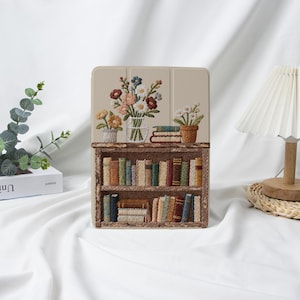 Puede incluir: Funda beige para tableta con un diseño bordado de una estantería con libros coloridos y jarrones con flores. La funda tiene un fondo neutro y una estética decorativa y vintage.