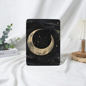 Puede incluir: Funda negra para tableta con diseño celestial. Presenta una gran luna creciente dorada y numerosas estrellas pequeñas sobre un fondo oscuro. El diseño evoca un tema de cielo nocturno, ideal para proteger una tableta.