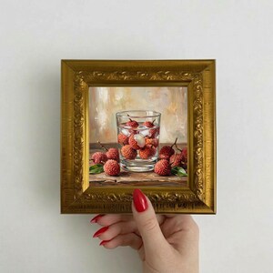 May include: A framed painting of a glass of water with ice and lychees, surrounded by more lychees. The painting is in a gold ornate frame. The background is a soft, blurred painting.