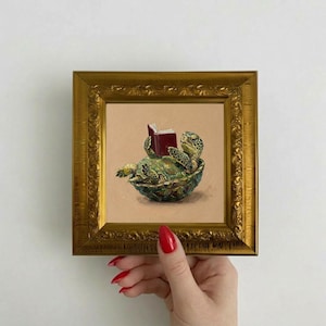 Puede incluir: Pintura enmarcada de una tortuga verde leyendo un libro rojo. La tortuga está en su caparazón, sobre un fondo beige. El marco dorado adornado añade un toque clásico a la obra.