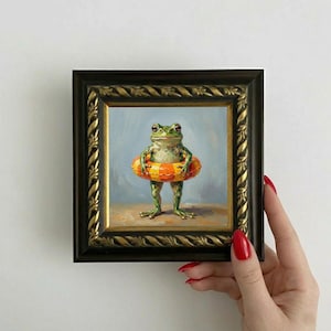 May include: A framed painting of a green frog wearing an orange and yellow inflatable ring. The frog is standing on a sandy surface against a light blue background. The frame is black with gold accents.
