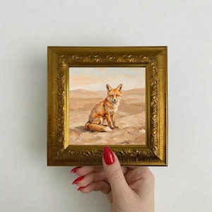 Könnte beinhalten: Gerahmtes Gemälde eines Fuchses in einer Wüstenlandschaft. Der Fuchs hat orangefarbenes Fell und sitzt auf einem sandigen Hügel. Das Gemälde befindet sich in einem goldenen, verzierten Rahmen. Der Hintergrund zeigt eine Wüstenlandschaft mit hellblauem Himmel.