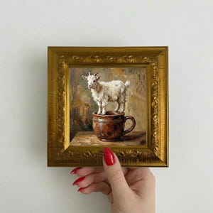 May include: A framed painting depicts a white goat standing in a brown mug. The artwork is set against a textured background with warm tones. The frame is gold-colored and ornate, held by a hand with red nail polish.
