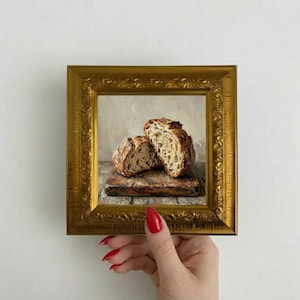 May include: A small square painting of two halves of a crusty sourdough loaf resting on a wooden board, enclosed in an ornate, metallic gold-colored frame with intricate embossed floral patterns along the edges and corners of the border.