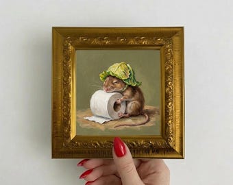 May include: A small oil painting in an ornate gold frame depicting a brown mouse wearing a green cabbage leaf as a hat. The mouse is resting its head on a white roll of toilet paper against a muted olive green background.