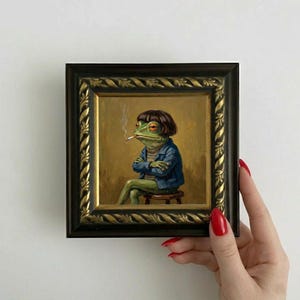 Pode incluir: Pintura emoldurada de um sapo antropomórfico com jaqueta jeans, fumando um cigarro e sentado em um banquinho. O sapo tem aparência humana e está em um fundo dourado. A moldura é escura com detalhes dourados.