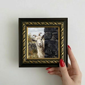 Könnte beinhalten: Gerahmtes Gemälde einer weißen Ziege, die hinter einer dunklen Steinmauer hervorschaut. Das Kunstwerk befindet sich in einem quadratischen, verzierten schwarzen und goldenen Rahmen. Die Ziege hat Hörner und einen freundlichen Ausdruck.